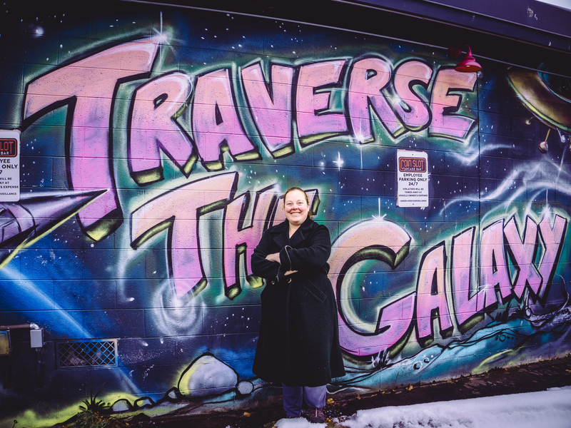 Photo of founder, Sabrina Jordan, in front of the Traverse The Galaxy mural outside of The Coin Slot in Traverse City, Michigan