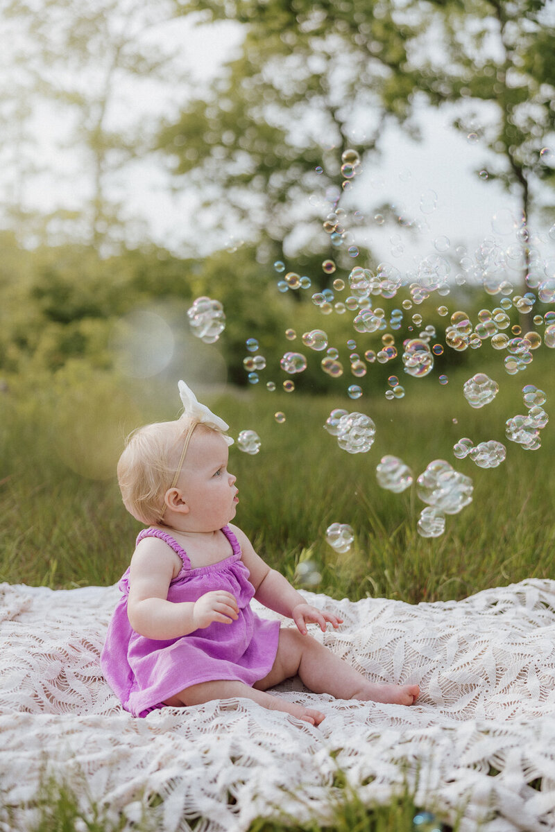 Baby sitting on a blanket with bubbles blowing during her first birthday portrait session in De Soto, Missouri. 