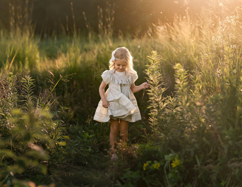 A little girl playing in the tall grass in a cream dress at sunset