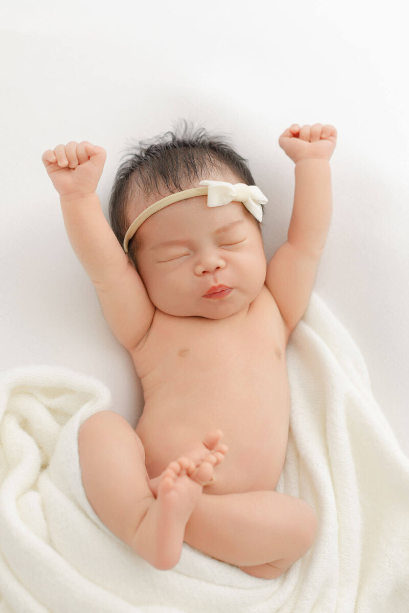 Newborn baby sleeping with arms raised up in the air laying on white cozy backdrop with white blanket wrapped around outside of body. Cozy newborn photo studio in Portland, Oregon.