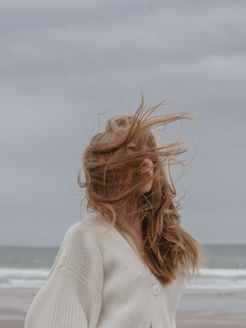 Langhaarige Frau in kuscheliger Strickjacke am Strand, das Gesicht vom Wind verdeckt, während die Haare in alle Richtungen wehen - ein Moment der  Selbstreflexion und des Aufbruchs