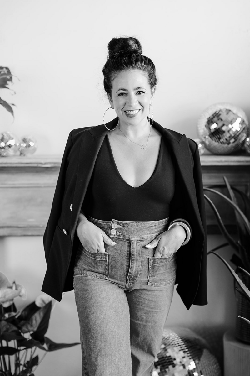 black and white portrait of woman in black tank top, high waisted jeans, and blazer over her shoulders. she has disco balls behind her