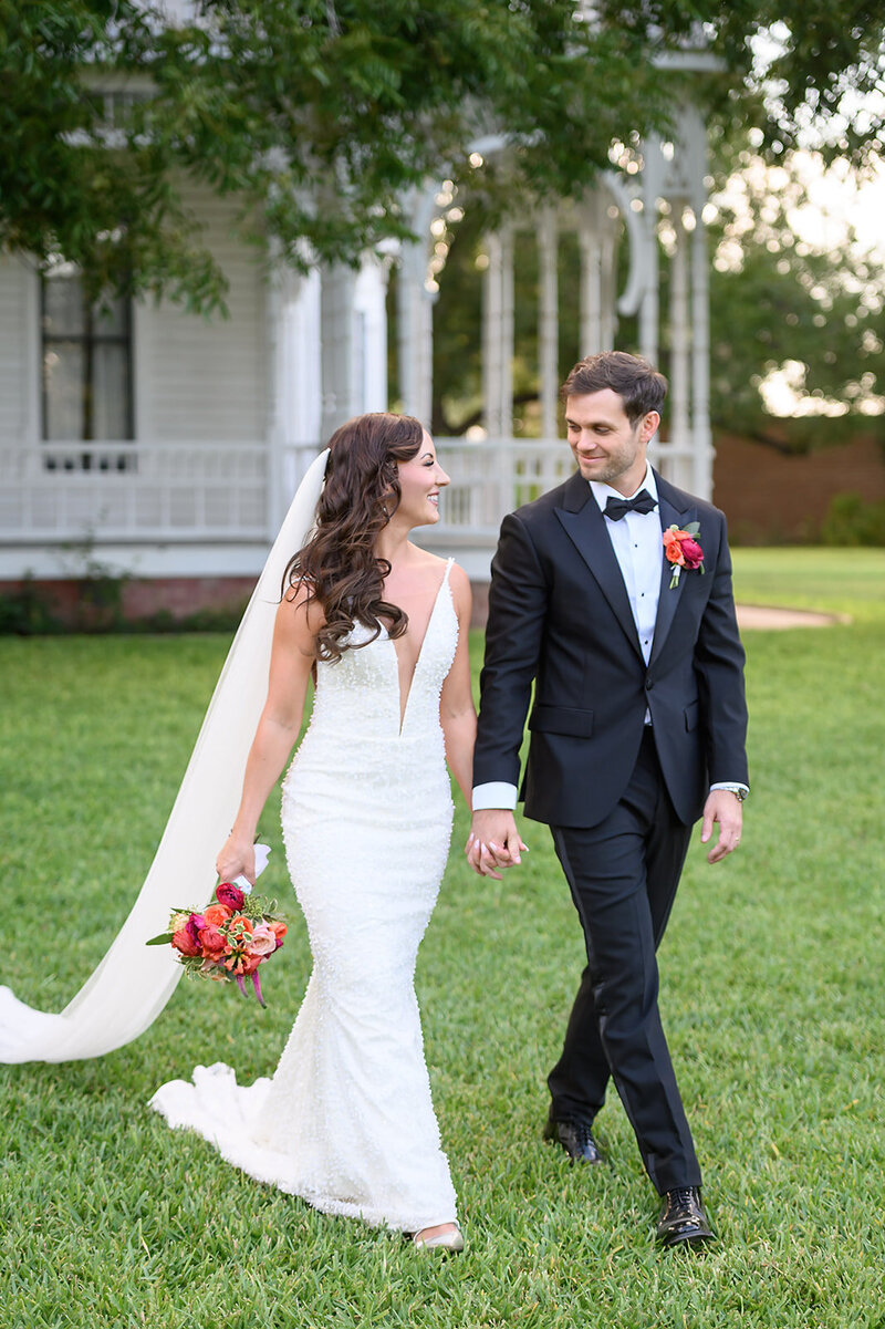 Newlyweds stroll hand-in-hand, bride holding a bright bouquet in rich pink and orange tones—colorful and romantic.