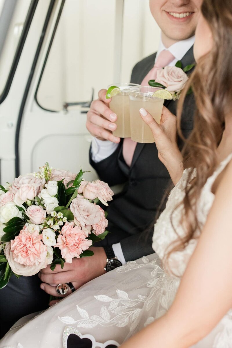 Close-up of a smiling bride and groom sitting in a vintage bar cart at the August House venue, each holding a cocktail and featuring a pink floral bouquet, capturing a joyful wedding moment at this Dickinson, North Dakota wedding venue