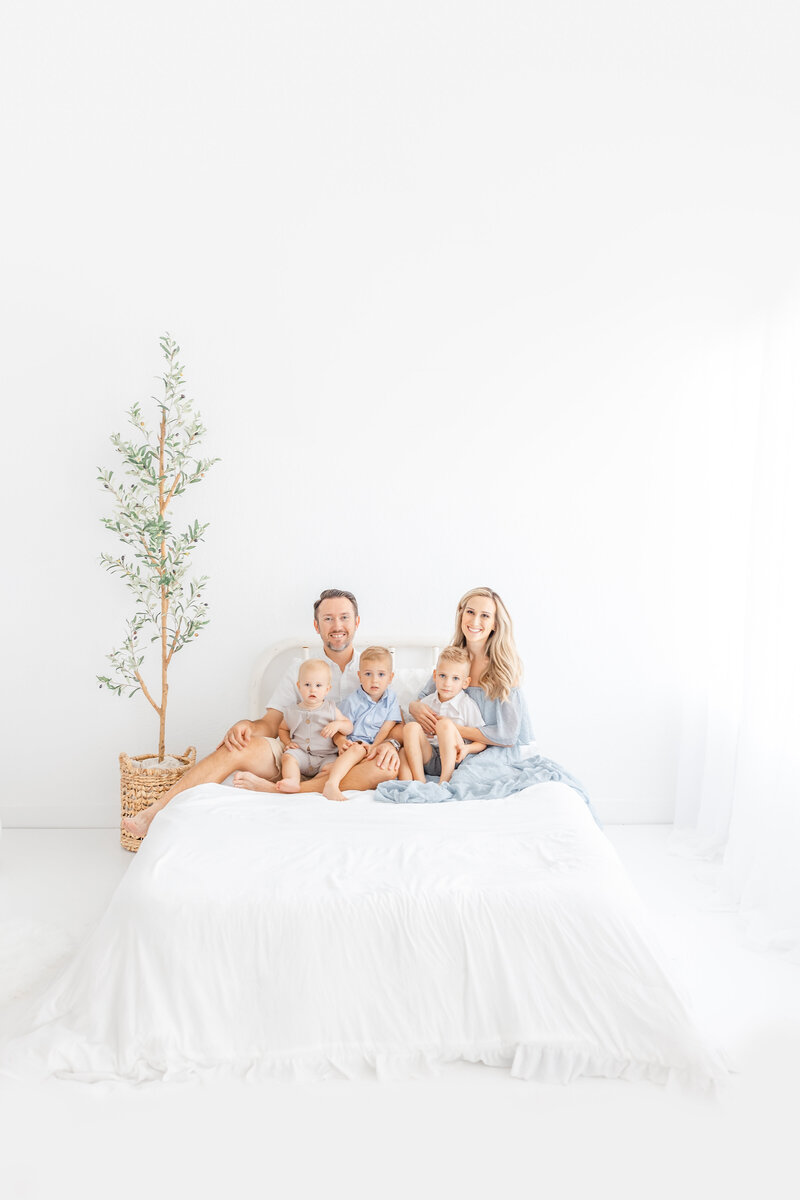 nichole pach in blue dress sitting on bed with her husband and three boys in white studio