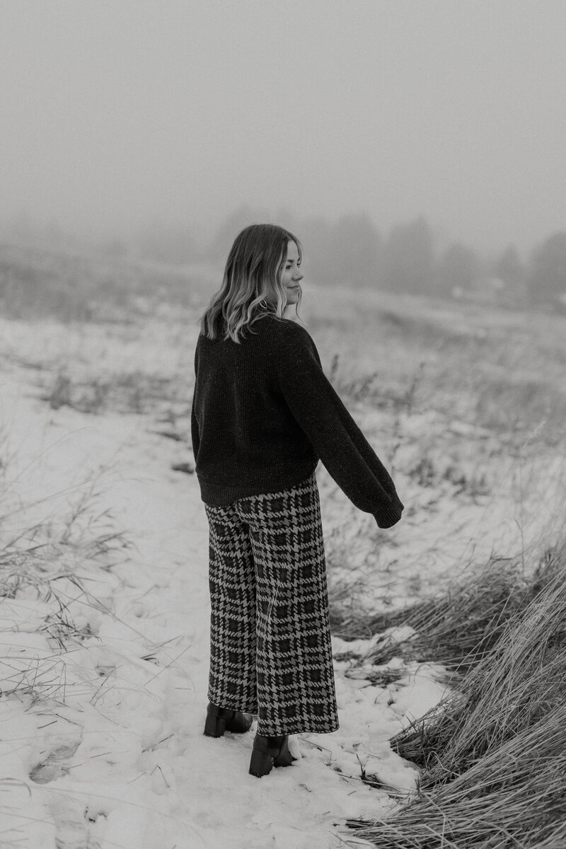 Black and white photo of a women wearing a dark sweater and funky checkered pants in the snow, her right profile shows her smiling slightly
