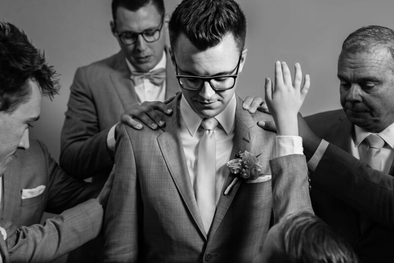 Groom surrounded by groomsmen while being prayed over. Little boy stretches to reach.