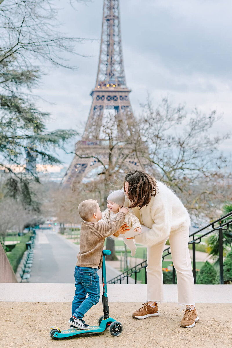 Mother lifting a baby up toward a young boy on a scooter in front of the Eiffel Tower at Trocadéro during a playful Paris family photoshoot.