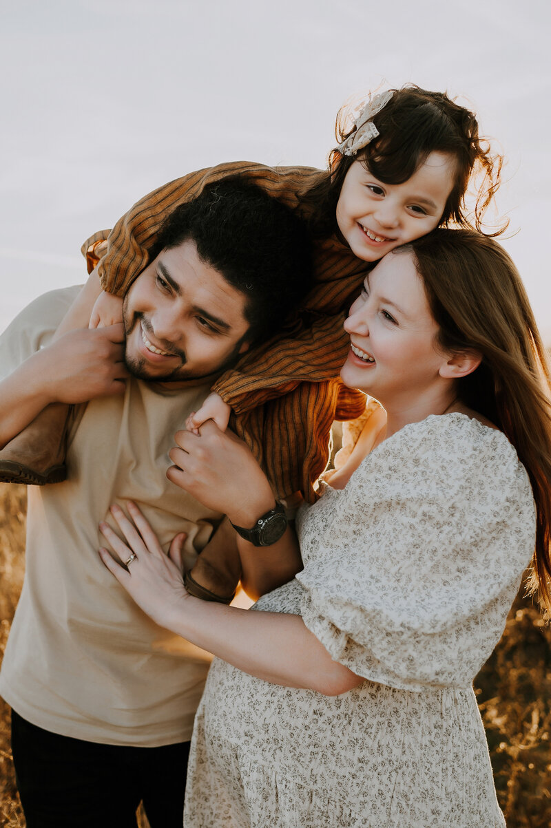 A dad holding his daughter on his shoulders while mom stands beside him looking up and smiling at Murrell Park in Flower Mound, Texas