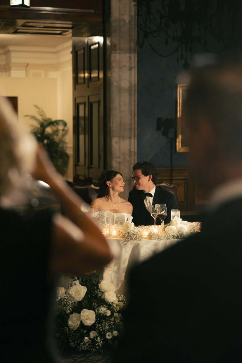 Woman and man in pantsuits stand in front of Chicago subway. Woman is holding a bouquet of roses while man kisses her on cheek