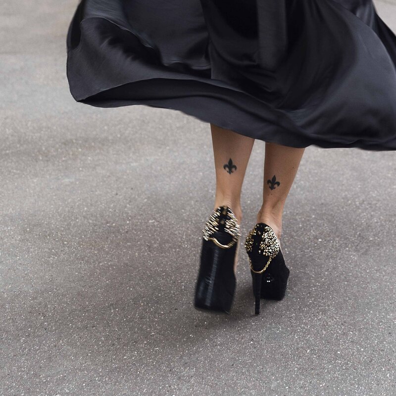 plan sur les pieds d'une femme avec des chaussures à très hauts talons et séquins, des tatouages de fleurs de lys sur l'arrière des chevilles et une robe noire