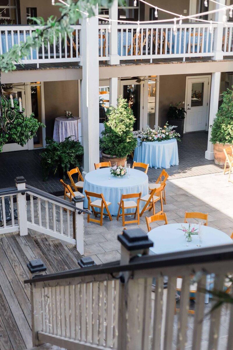 An outdoor courtyard set for an event, with round tables covered in white cloths, wooden chairs, floral centerpieces, potted plants, and string lights overhead.