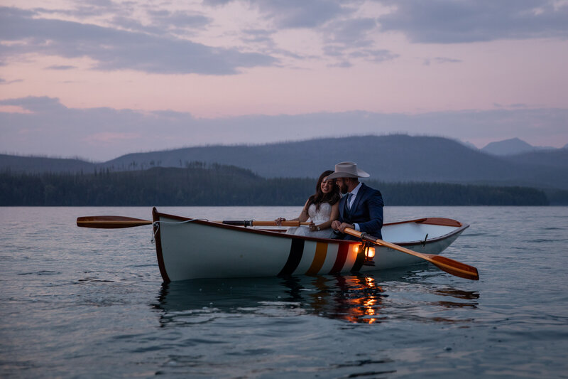Bride and groom row across a tranquil Montana lake at sunset in a vintage wooden canoe with a lantern glowing at the bow. The bride wears a white wedding dress, and the groom, dressed in a blue suit and cowboy hat, smiles as they enjoy their adventure elopement in the mountains.