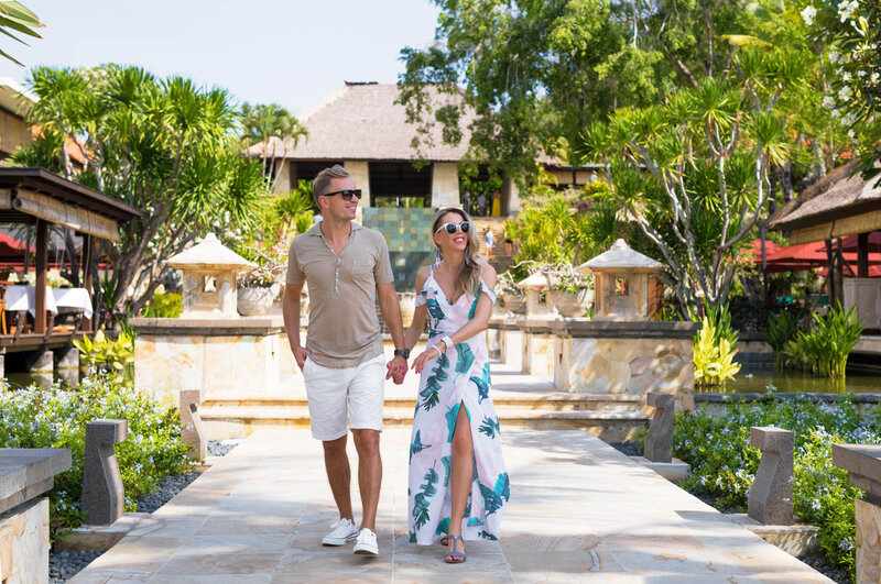 Couple holding hands and walking through a tropical resort garden with palm trees and stone pathways.