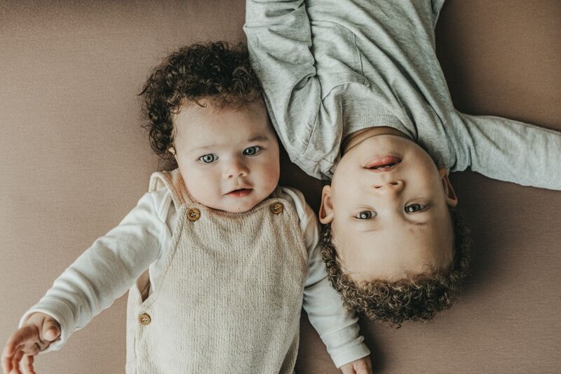 two adorable siblings with curly hair lying side-by-side on a soft brown surface, looking up at the camera with curious expressions by Ruaa photography