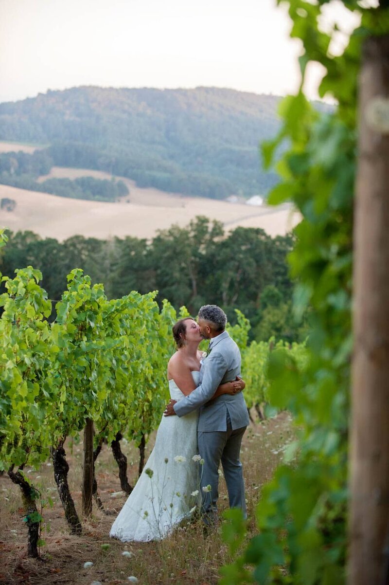 a bride and groom kiss between rows of grape vines