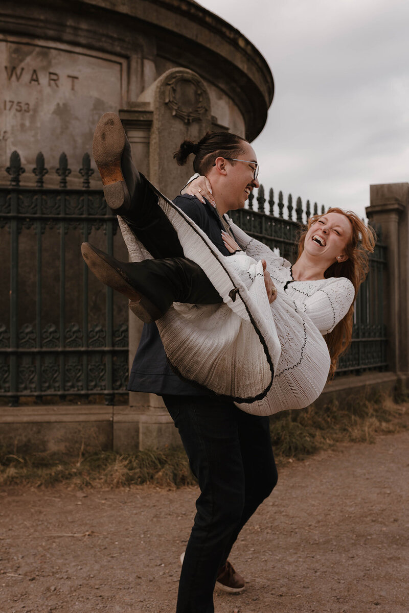 Playful couple’s portrait in Edinburgh’s Old Town, with the groom lifting his partner in front of an historic Stewart tomb, captured in a cinematic fine art style by Scottish wedding photographer Aly Robinson.