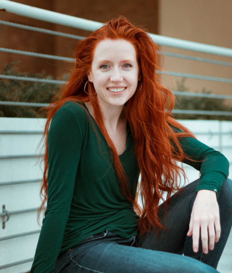 Sage is a fair skinned smiling young woman with long red hair and blue eyes. She is wearing a long sleeved vneck shirt that is blue green.