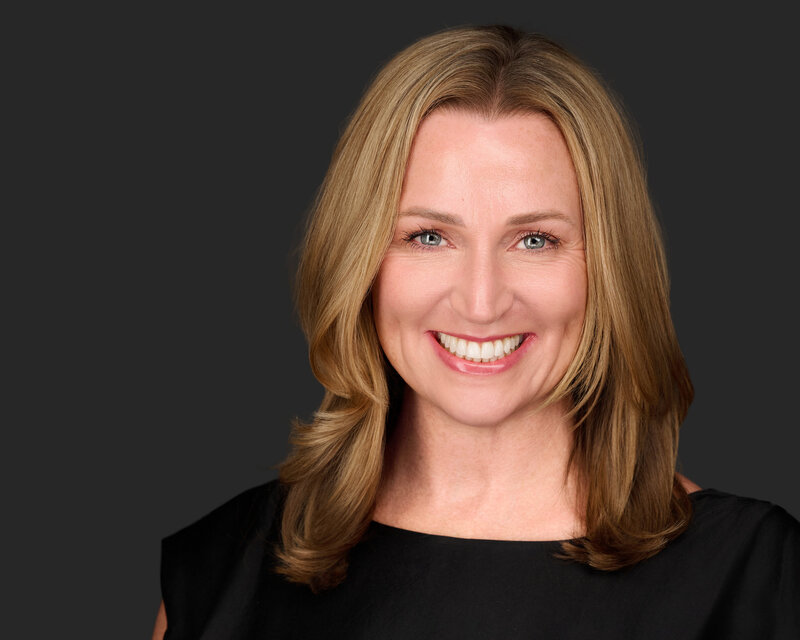 Jenn Cloutier, self-leadership coach, smiling warmly with shoulder-length blonde hair, wearing a black professional top against a dark gray background.