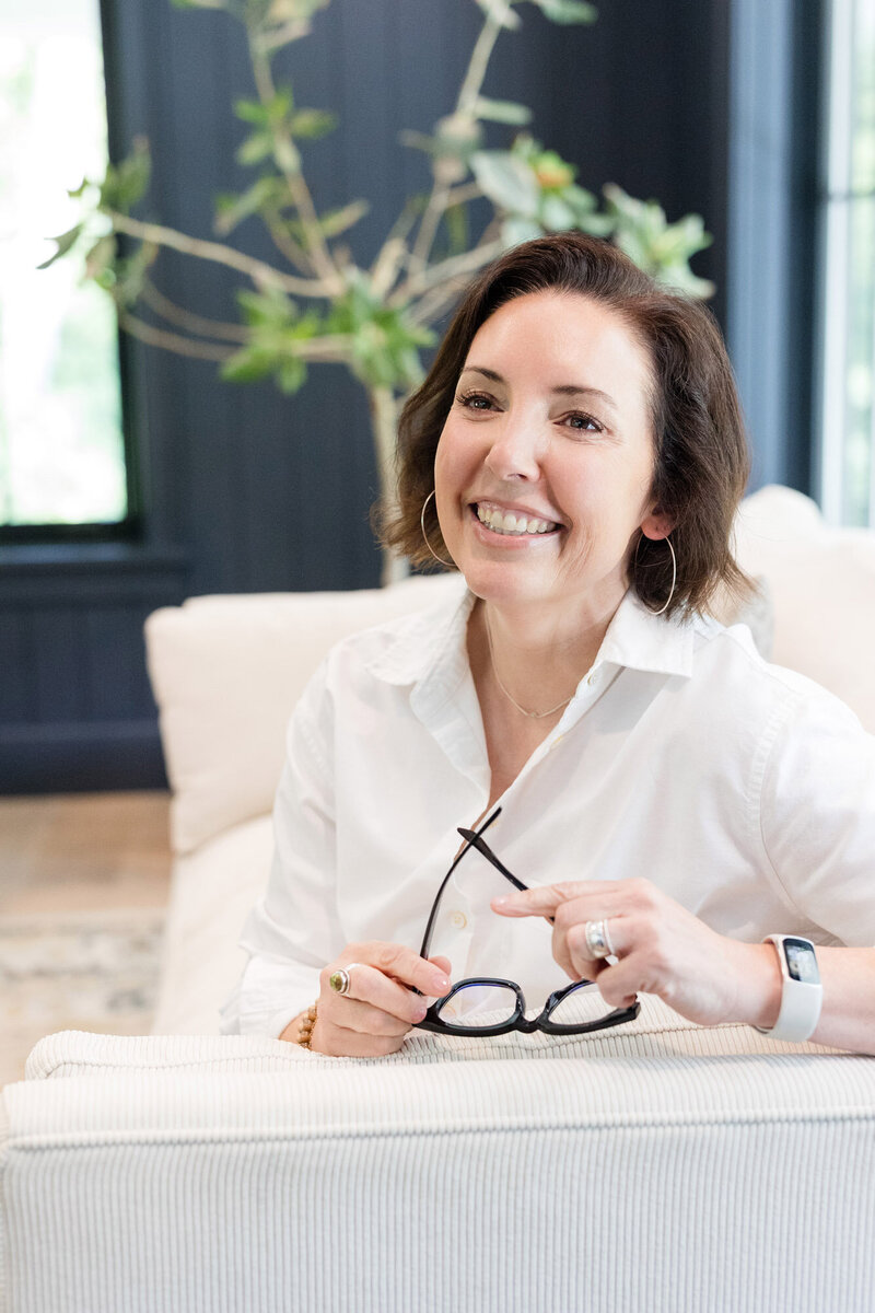 Life coach Dinah McBridge sitting on a couch, holding a pair of glasses in her hands, smiling