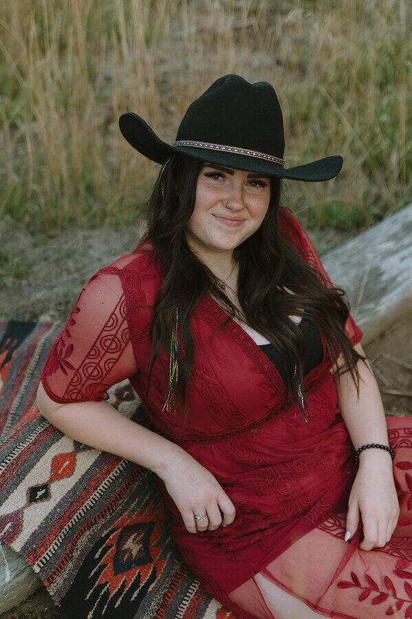 Close up portrait of a woman wearing a red lace dress and a black cowgirl hat. She's leaning against a log that's been covered with a woven blanket.