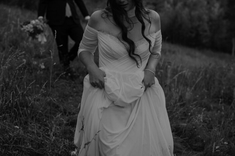 Bride walks through mountain field holding her dress.