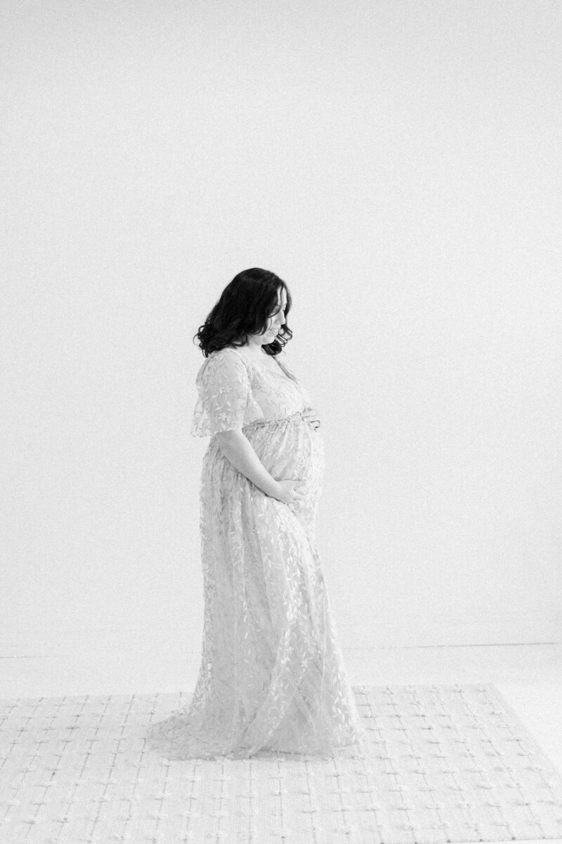 Serene black and white maternity portrait of mother in lace evening gown looking softly down and gently holding her baby bump in a neutral studio by Cambridge, MA maternity photographer Fieldstone Studio