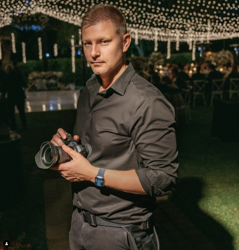 A professional LA wedding photographer in an all-black outfit holding a camera at a beautifully lit outdoor wedding reception. String lights illuminate the background, creating a romantic ambiance for a luxury wedding celebration.