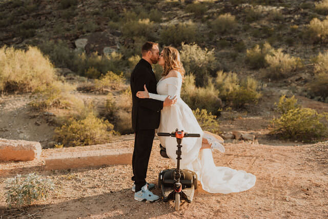 A bride and groom kissing while the bride is kneeling on a scooter.