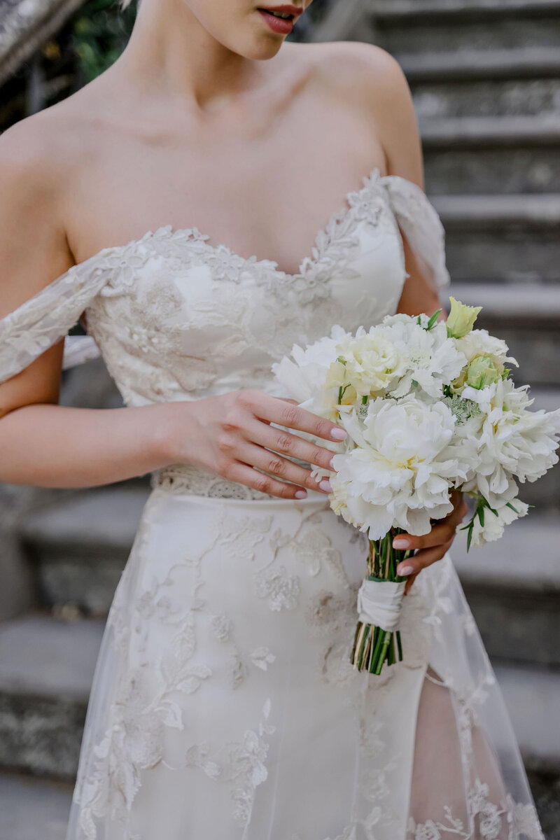 A close-up of a bride in an elegant lace wedding gown holding a white floral bouquet featuring peonies and greenery, standing on a stone staircase