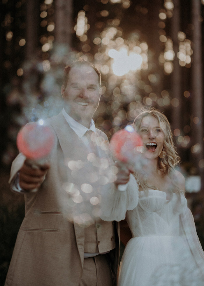 A wedding couple pointing and shooting bubble toy guns at the camera and smiling and laughing.