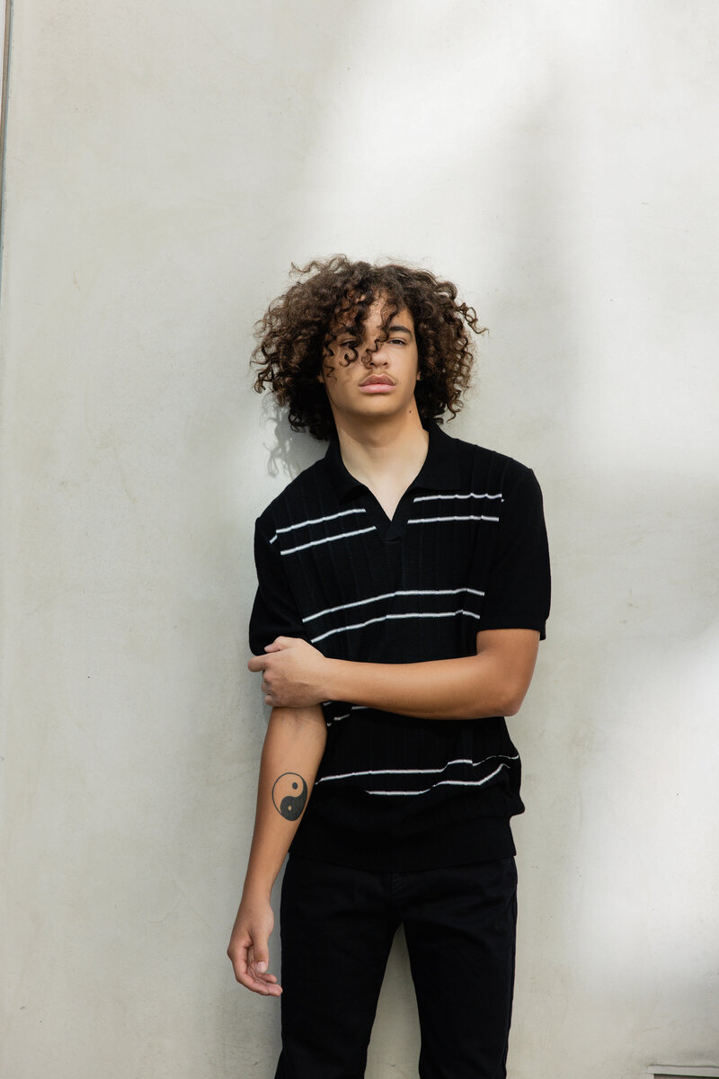 Teen boy with curly hair wearing a black short-sleeve sweater with white stripes, standing confidently against a clean white backdrop with arms folded.