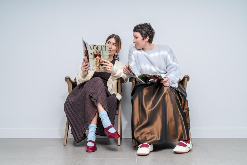 Two wardrobe stylists sitting in chairs in stylish outfits reading Vogue magazine against neutral background