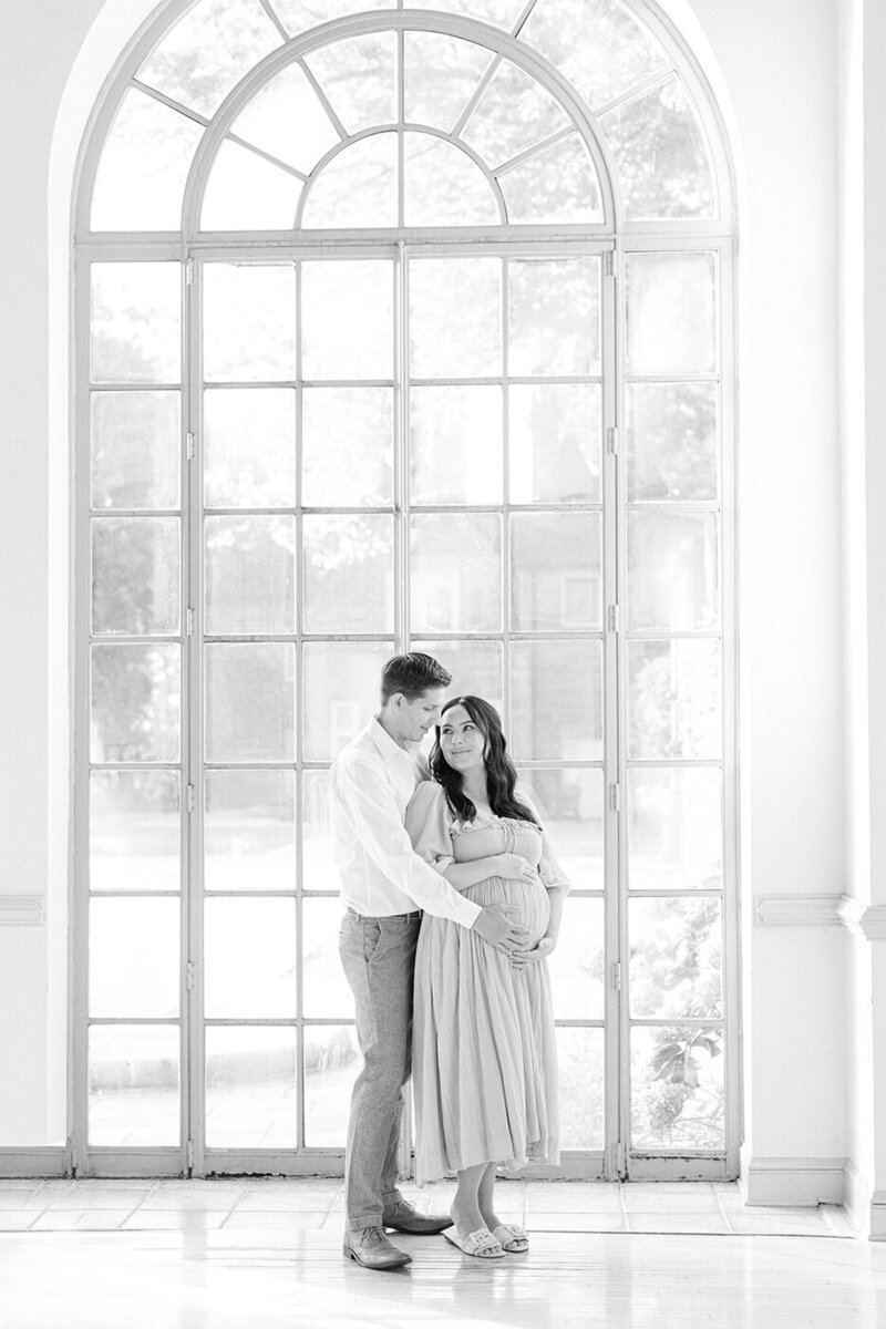 pregnant couple holding belly standing in front of large glass window by philadelphia newborn photographer