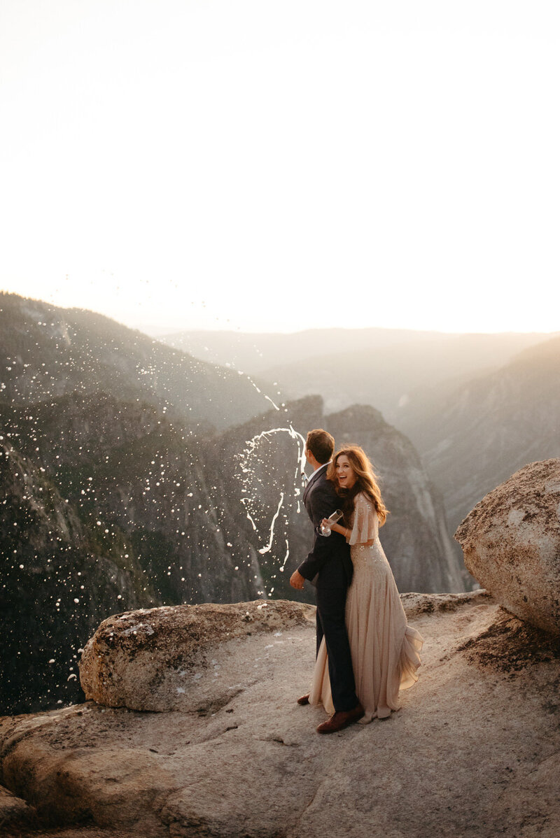 Bride and groom pop champagne on the edge of a cliff after they got their Yosemite Wedding Permit 