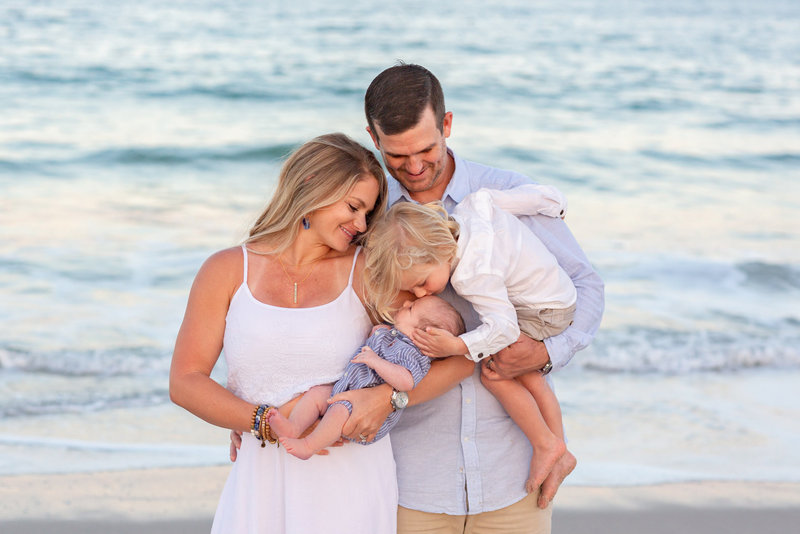 parents on the beach wearing white and blue holding a toddler kissing a newborn  Johns Island Moorings