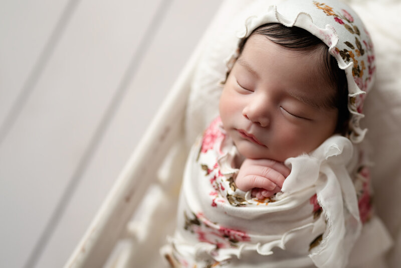 Timeless close-up of a newborn girl resting in a white crib, adorned in a floral bonnet and heirloom wrap.