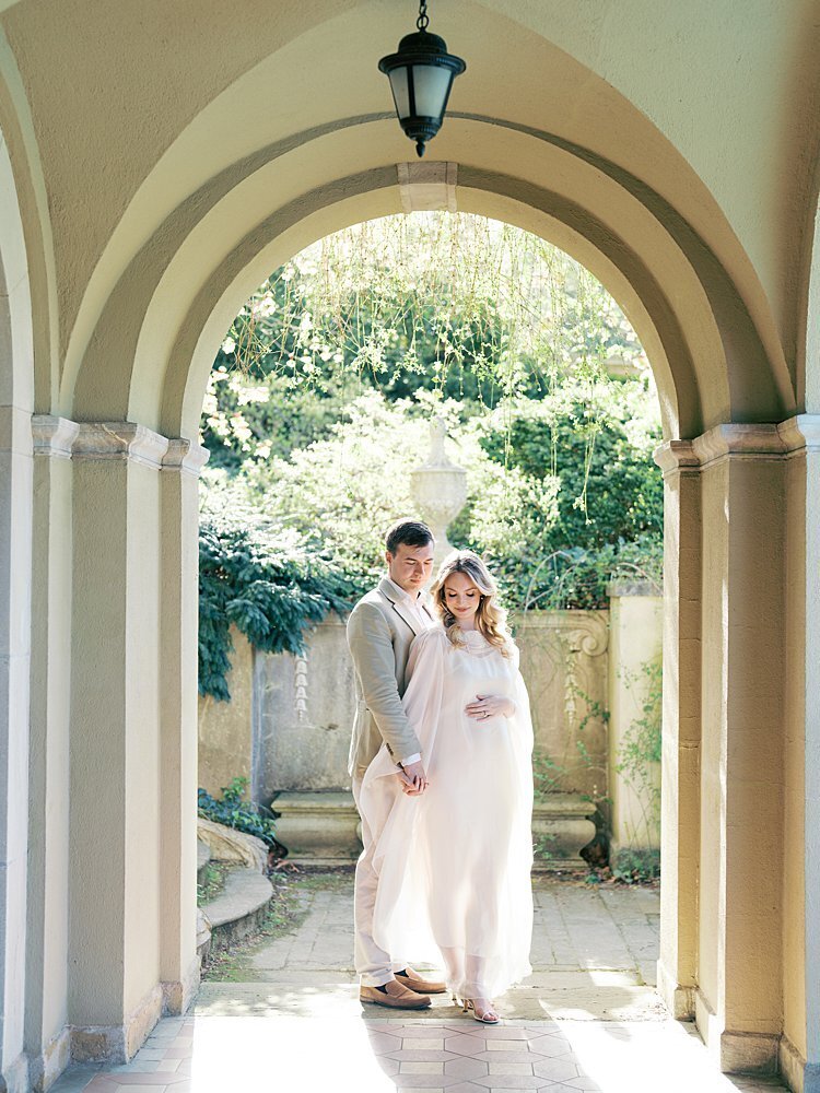 An expecting couple stand together in an archway at Dumbarton Oaks Gardens in DC during their maternity photos.