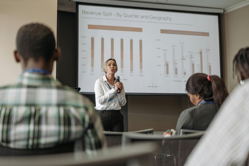 White middle aged woman speaks to a group during a meeting/training session. Behind her is a projector screen with two graphs discussing revenue splits by quarter and geography