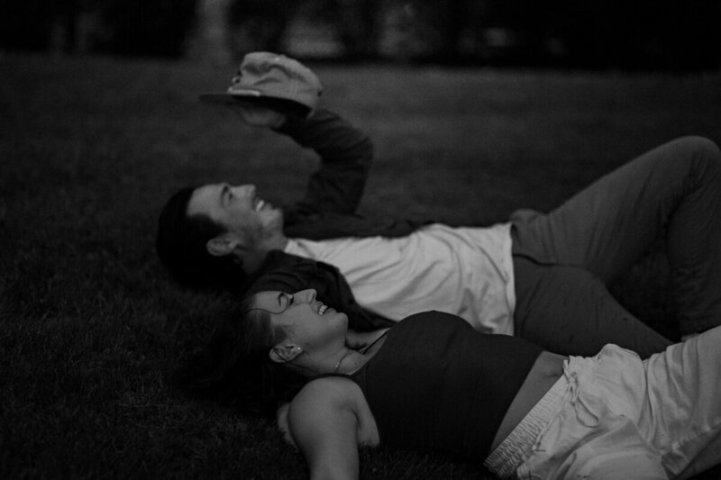 A black and white image captures the essence of a New England elopement: two people lying on grass at night, one holding a hat up. Relaxed and happy, they gaze upwards, with trees faintly visible behind them. The scene feels like it's been framed through the lens of a New England photography wedding artist.