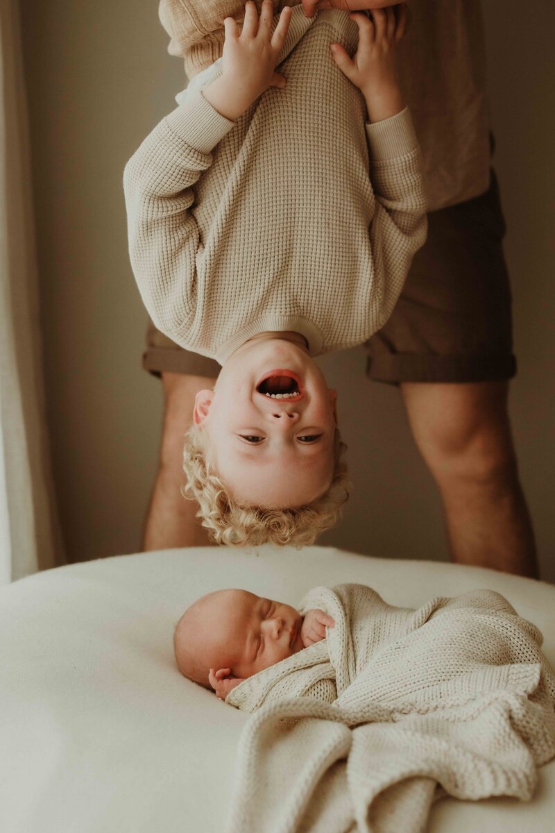 Playful child hanging upside down while newborn sleeps peacefully, photographed by Sydney family and newborn photographer Sophie Lea.
