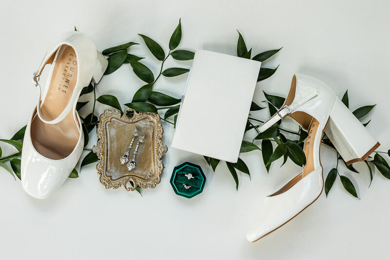 Flat lay of shoes, earrings, rings and book of vows with greenery