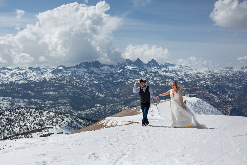 Groom leads his wife along a snow covered mountaintop. He holds his cowboy hat on his head hiding from the wind, her dress and hair blowing in the wind. Jagged mountains line the background.