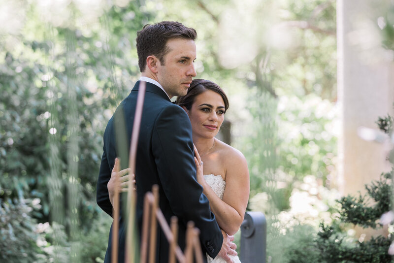 white groom in a black tux with his back to the camera holding a white brunette bride in a forest both looking in the distance