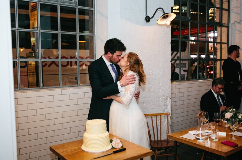 Sweet celebration: Bride and groom kiss near cake at Clissold House reception.