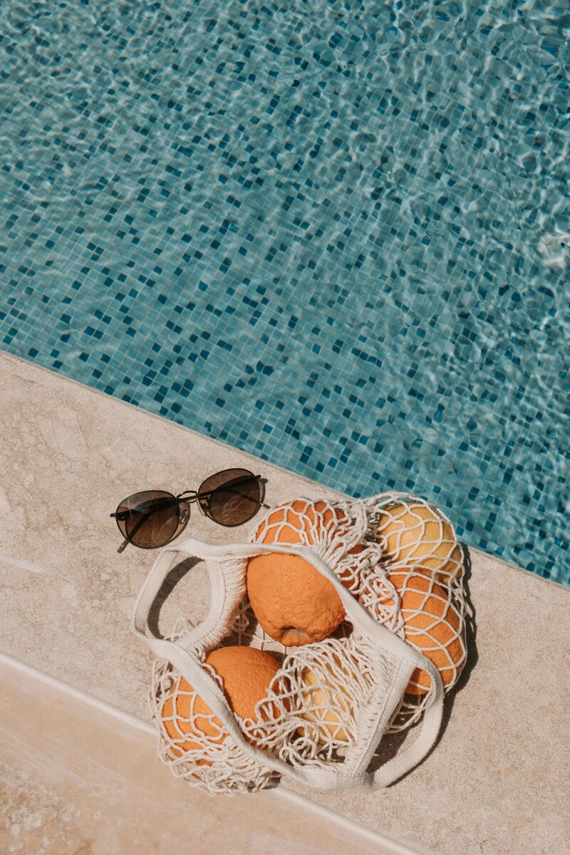 A mesh bag filled with vibrant oranges rests on a sunlit stone poolside next to a stylish pair of sunglasses. The shimmering blue tiles of the pool add a refreshing backdrop, symbolizing the luxurious yet relaxed experiences that vacation fundraising for charities can provide while supporting meaningful causes.