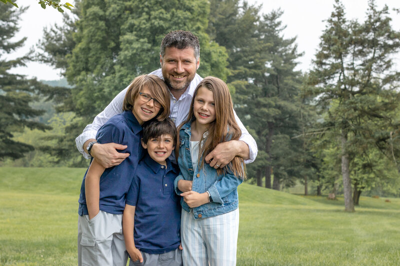 A candid outdoor portrait of a smiling mother holding her young son in a park, showcasing natural light and relaxed interaction, captured by a Delaware family photographer. The mother is looking lovingly at her son, and both are dressed in light colors with greenery in the background, highlighting a warm family moment