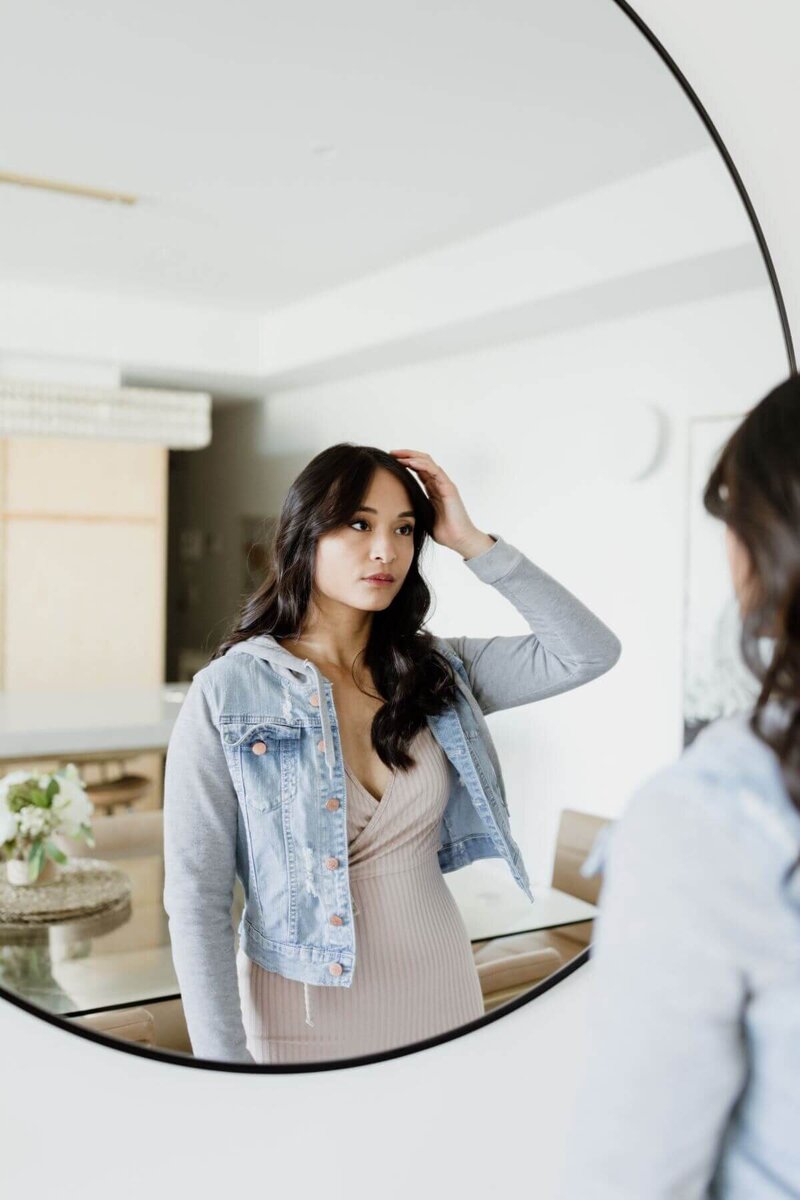 Trang Nova looking at her reflection in a mirror, fixing her hair. She is wearing a beige ribbed dress and a denim jacket. 