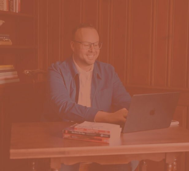 Dr. Mike Stokes smiling while working on a laptop in a wood-paneled office, warm orange overlay applied.