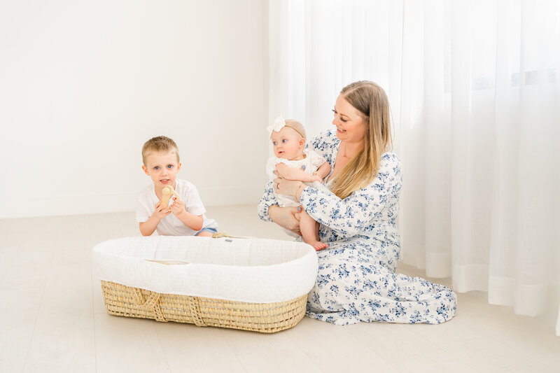 a mother and her toddler boy and baby girl sit on the floor of a white photo studio in Austin, TX. In front of them is a Moses basket. The mother wears a long floral blue dress. Her children wear a white outfit.